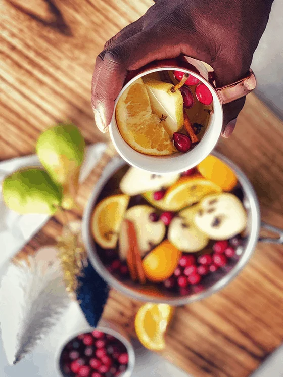 A woman holds a cup of pear cider with fruit slices over a pot of more of the cider.