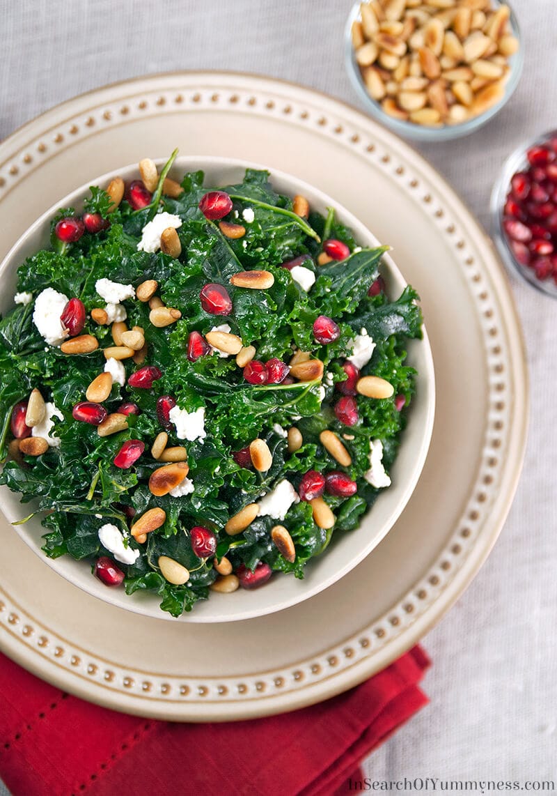 Massaged kale salad with white cheese, nuts, and pomegranate arils in a white bowl.