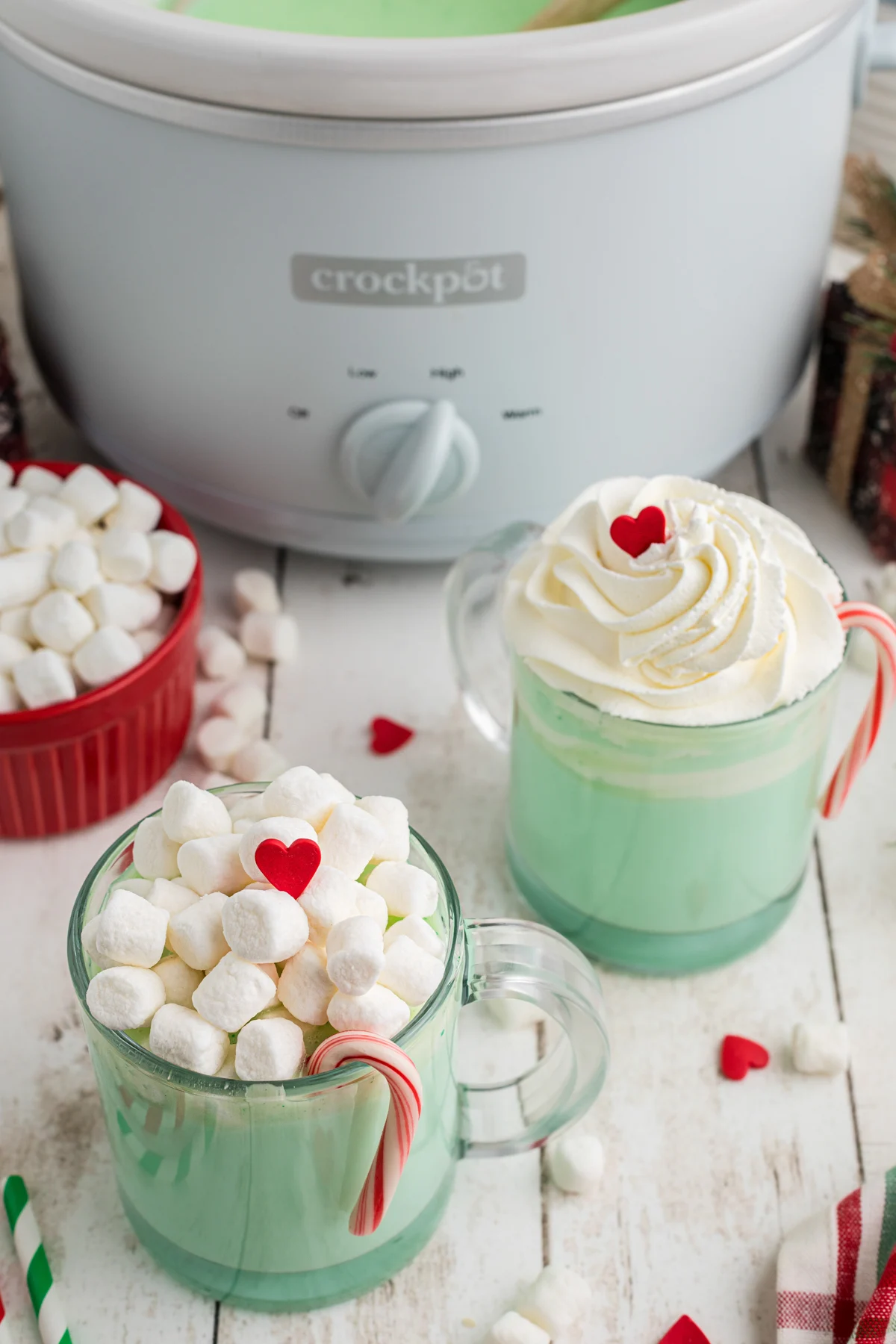 Grinch hot chocolate in two clear mugs with marshmallows on top and tiny red heart candies.