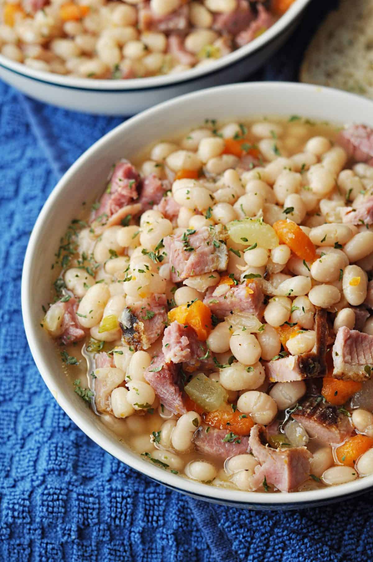 Ham and bean soup with carrots in two cream bowls next to a royal blue kitchen towel.
