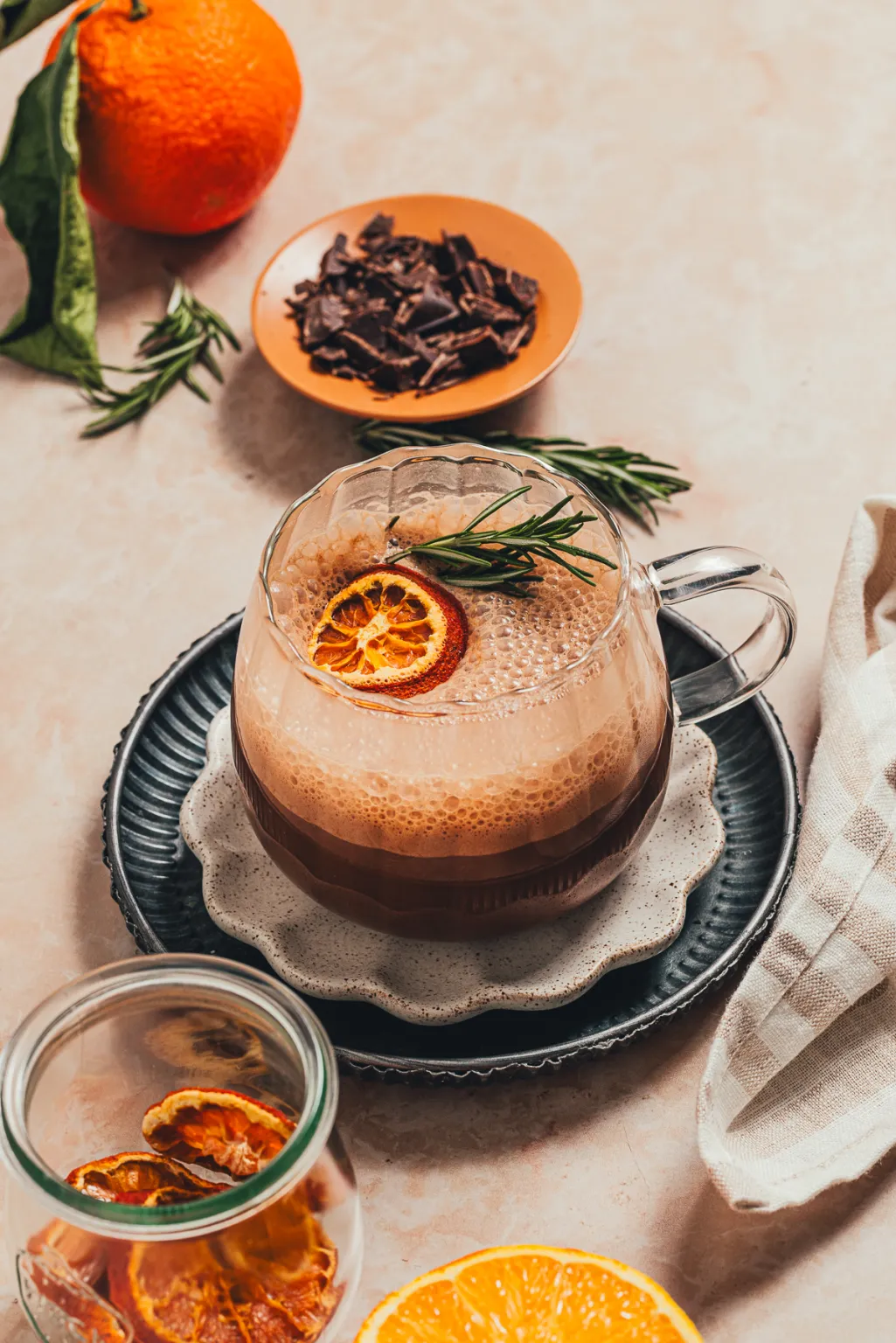 Rosemary orange hot chocolate in a clear glass with dried orange and rosemary garnish on stacked decorative plates.