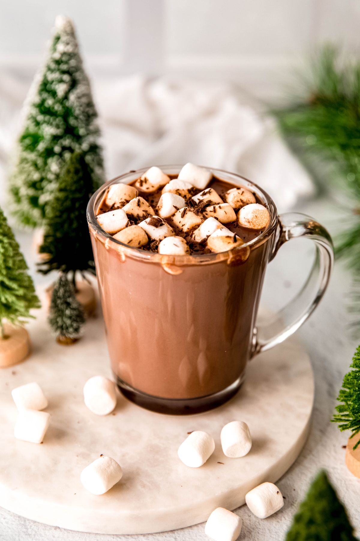 Dark hot chocolate in a glass mug with marshmallows on top on a wooden serving platter with a faux mini winter tree decoration.