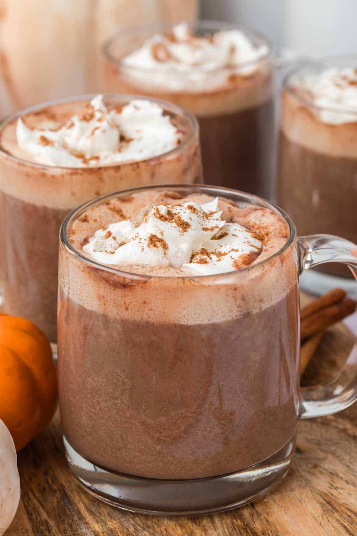 4 clear glasses of pumpkin spice hot chocolate with whipped cream and cinnamon on top.