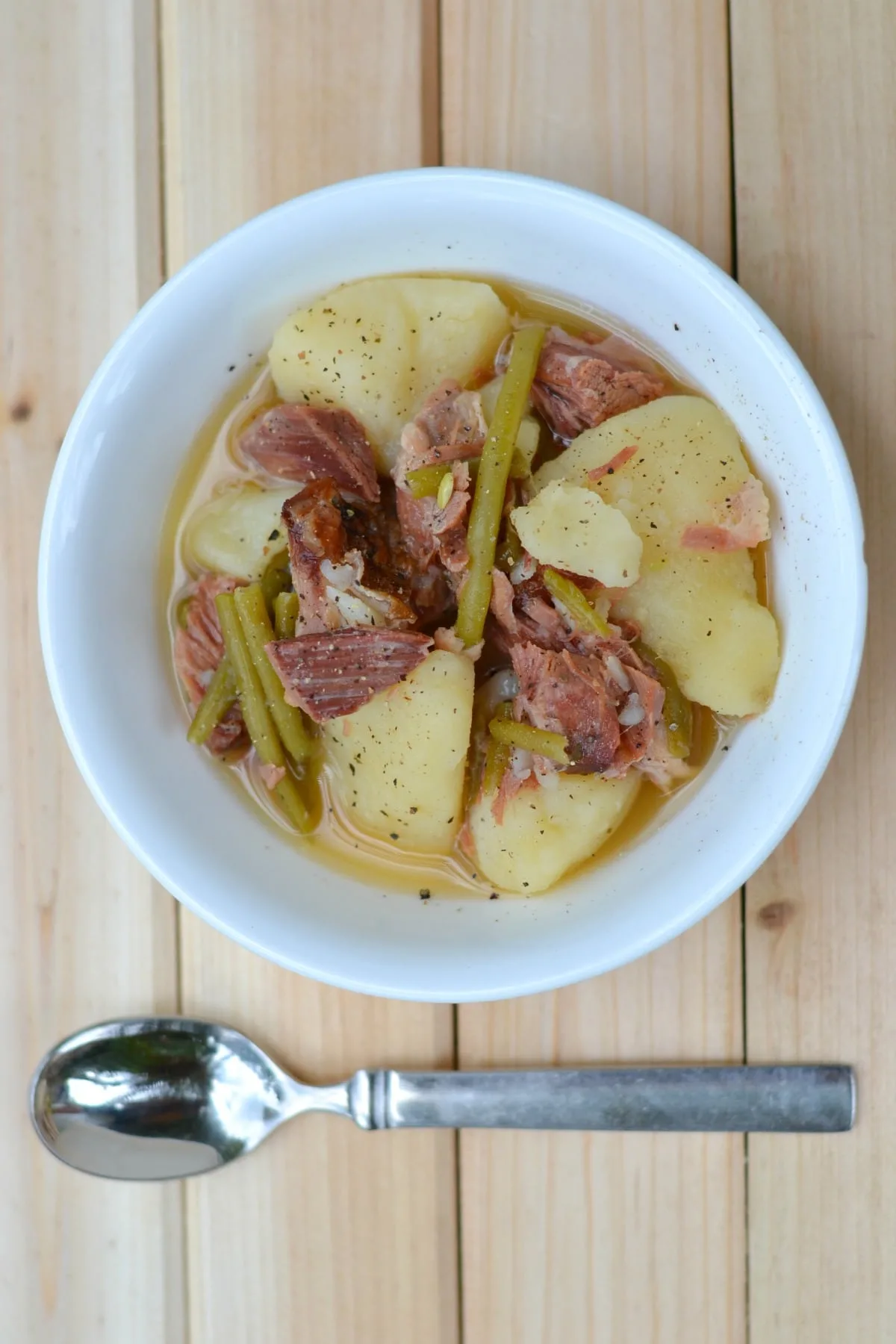 Ham, potatoes, and green beans in a white bowl next to a metal spoon.