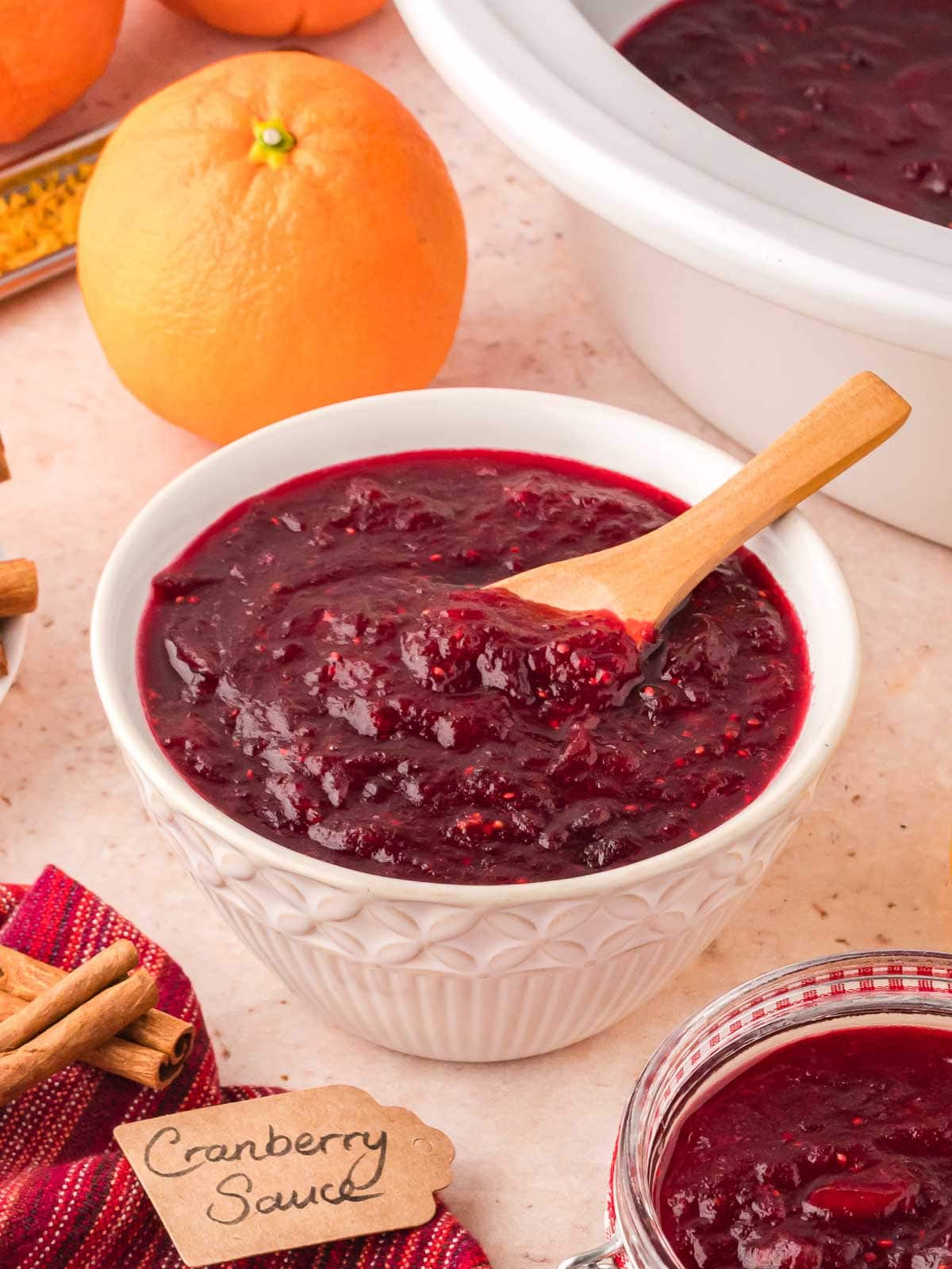 Crockpot cranberry sauce in a small white serving bowl with a mini wooden spoon in front of a white crockpot.