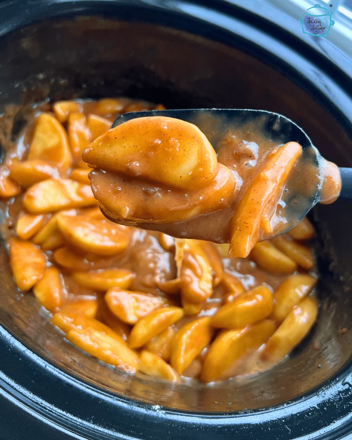 Crockpot apple pie filling in a black crockpot and lifted from the pot on a black spoon.