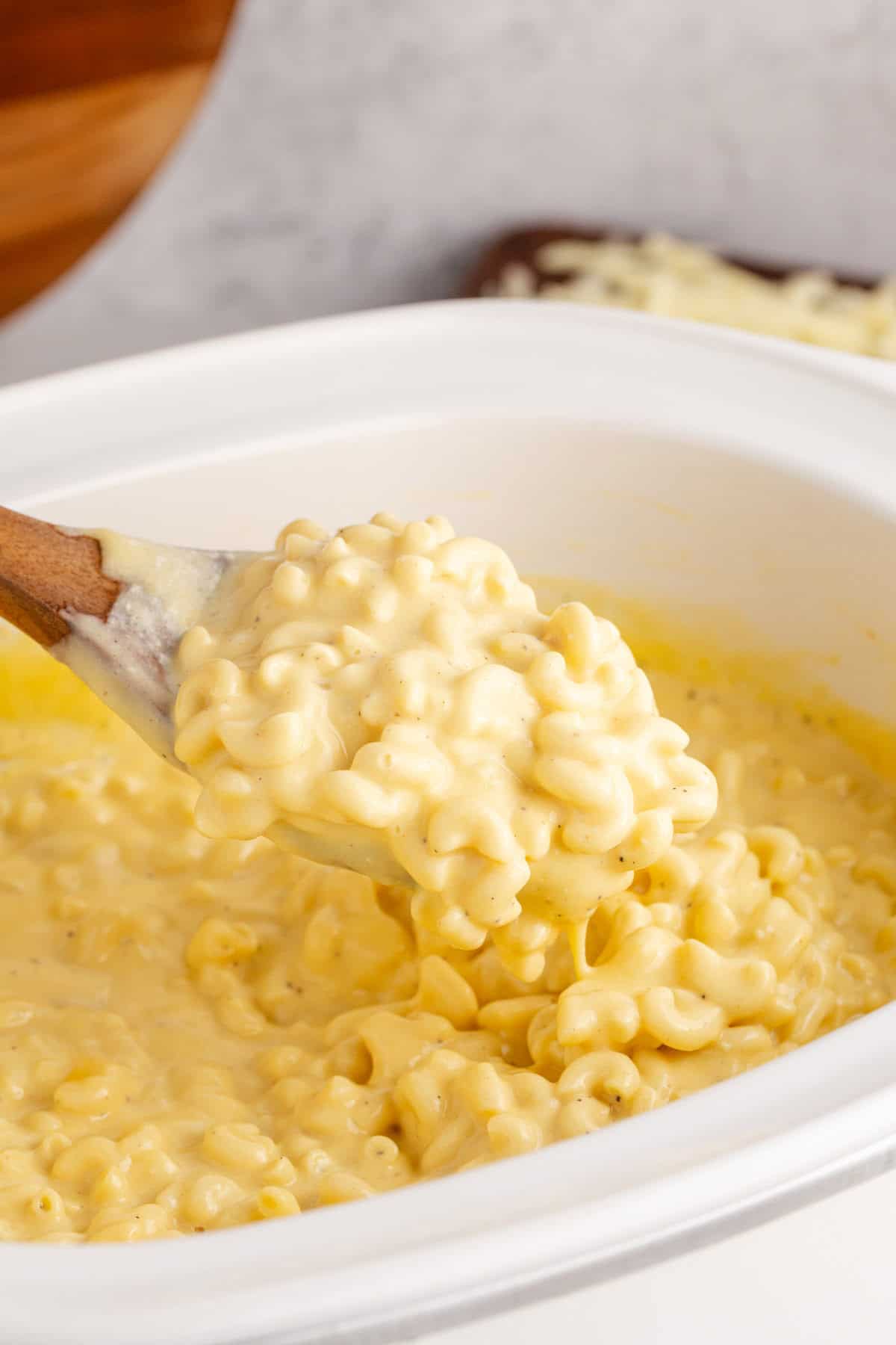Creamy yellow mac and cheese in a white crockpot and lifted from the pot on a wooden spoon.