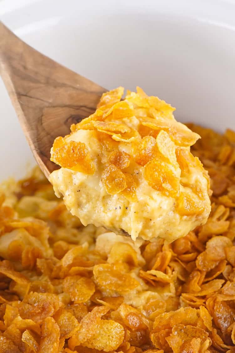 Funeral potatoes in a white crockpot and lifted from the pot on a wooden spoon.