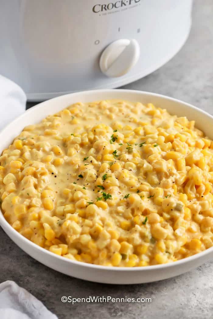Creamed corn in a shallow white bowl in front of a small white crockpot.