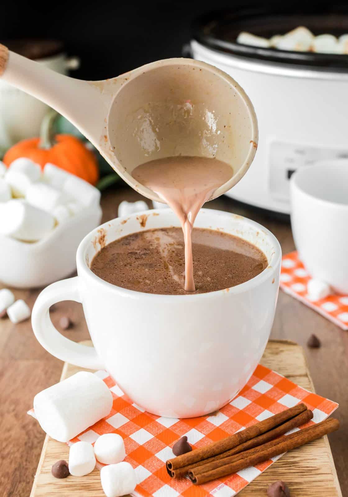 Pumpkin hot chocolate being ladled into a white mug on an orange and white checkered napkin.